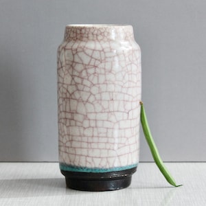 Può includere: Un vaso cilindrico in ceramica caratterizzato da una finitura smaltata bianca crepata con venature marrone scuro. La base del vaso è di colore marrone scuro uniforme, separata dal corpo superiore da una sottile fascia color ottanio. Un fagiolino verde poggia sul lato.