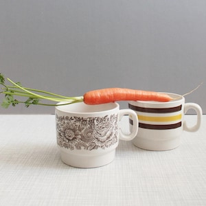Peut inclure: Deux tasses en céramique, l'une avec un motif floral et l'autre avec des rayures marron, jaune et blanc. Une carotte fraîche avec des feuilles vertes repose sur les tasses, créant une nature morte.