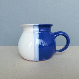 以下が含まれることがあります： 丸みを帯びたボディと大きなハンドルが付いた手作りのセラミックマグ。マグカップは垂直に分割され、半分は白、もう半分は鮮やかな青色です。内側とハンドルは青色です。マグカップの高さは約10cmです。