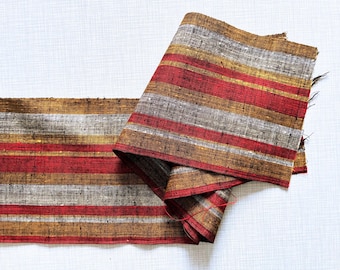 May include: A length of fabric with horizontal stripes in red, gray, and gold. The fabric is folded over on itself, revealing the striped pattern. The fabric is lying on a white surface.