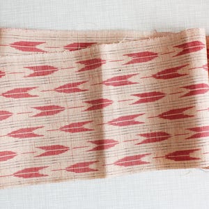 Op de afbeelding: Close-up van een gevouwen stof met een herhalend patroon van rode pijlvormige vormen op een beige achtergrond. De stof heeft een rafelige rand en een geweven textuur, wat duidt op een textiel voor knutselen of woondecoratie.