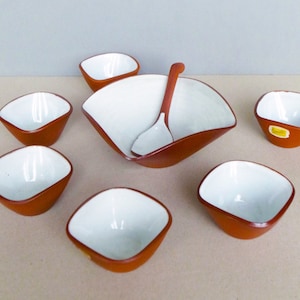 以下が含まれることがあります： 白の内側を持つテラコッタ色のセラミックボウル7個のセット。セットには、マッチするスプーンが付いた大きなボウルが含まれています。ボウルは茶色の表面に配置されています。