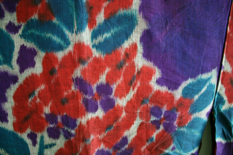 Op de afbeelding: Een close-up van een stof met een kleurrijk bloemenpatroon. De stof is een mix van rood, blauw, paars en wit.