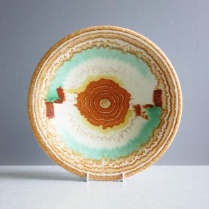 May include: A large, round ceramic bowl with a brown, green, and white abstract design. The bowl has a raised rim with a textured pattern.