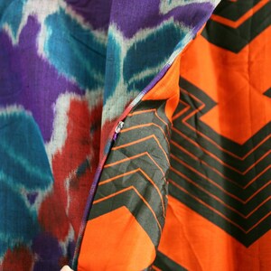 Op de afbeelding: Een close-up van een stof met een paars, blauw en rood ikat-patroon aan de ene kant en een feloranje stof met een zwart geometrisch patroon aan de andere kant.