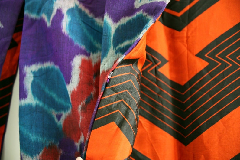 Op de afbeelding: Een close-up van een stof met een paars, blauw en rood ikat-patroon aan de ene kant en een feloranje stof met een zwart geometrisch patroon aan de andere kant.