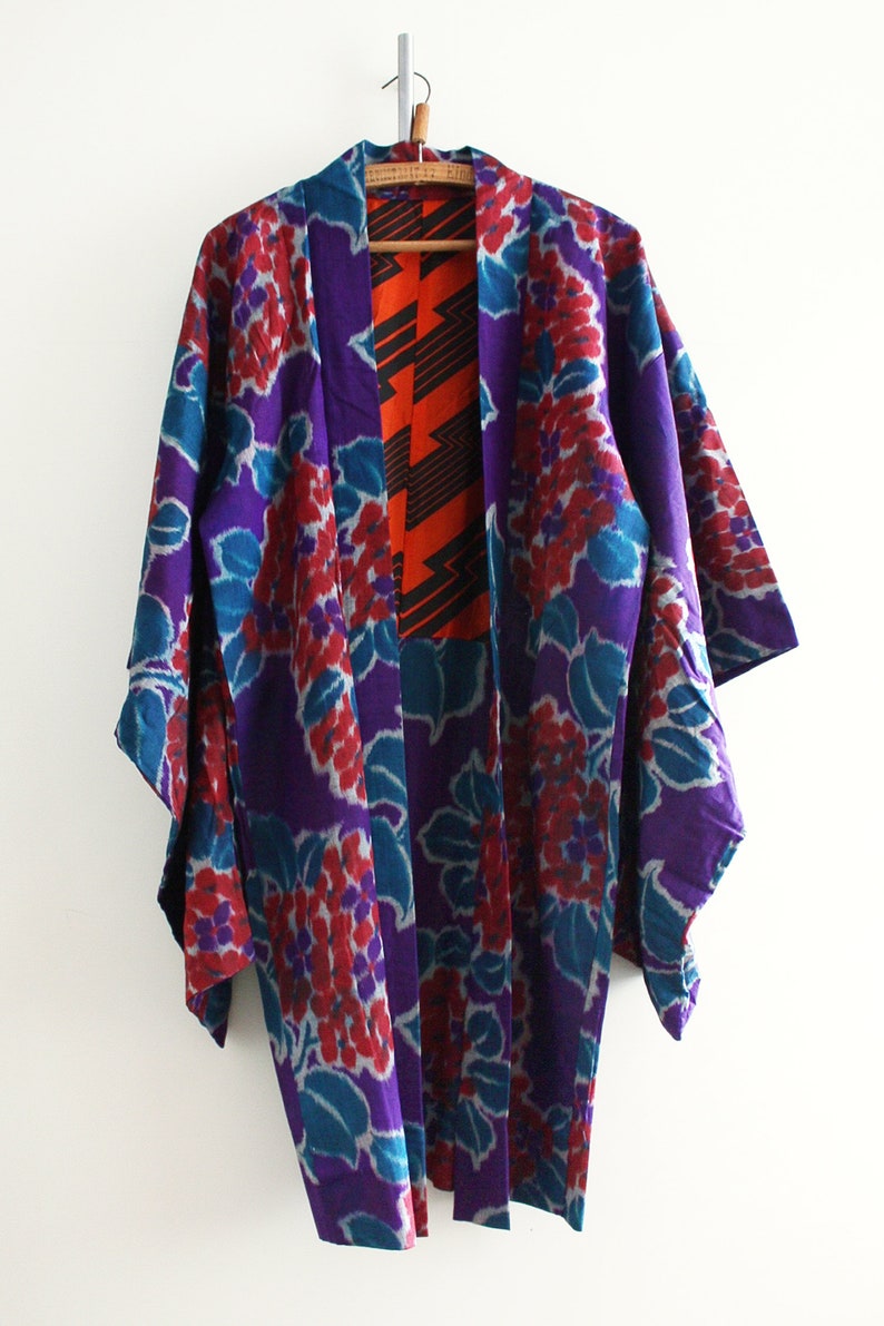 Op de afbeelding: Een paarse en turquoise kimono met een bloemenpatroon. De kimono heeft een rode en zwarte geometrische voering.