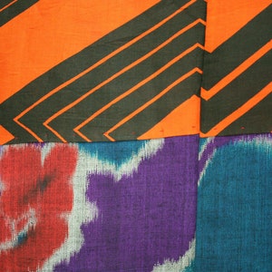 Op de afbeelding: Een close-up van een stof met een patchwork-ontwerp. Het bovenste gedeelte is oranje met zwarte diagonale strepen. Het onderste gedeelte is een paars en blauw ikat-patroon.