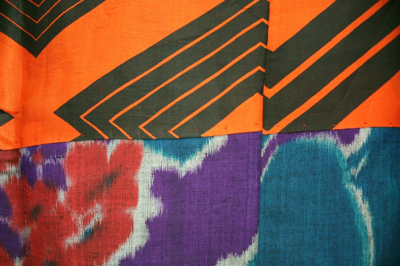 Op de afbeelding: Een close-up van een stof met een patchwork-ontwerp. Het bovenste gedeelte is oranje met zwarte diagonale strepen. Het onderste gedeelte is een paars en blauw ikat-patroon.