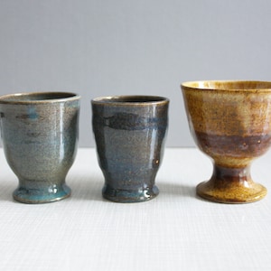 Op de afbeelding: Drie keramische bekers met verschillende glazuren. De beker links is blauw en bruin, de beker in het midden is donkerblauw en bruin, en de beker rechts is bruin en geel met een voet.