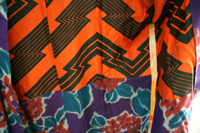 Op de afbeelding: Een kleurrijke stof met een geometrisch patroon in zwart en oranje. De stof is aan elkaar genaaid met een paarse en blauwe bloemenstof.