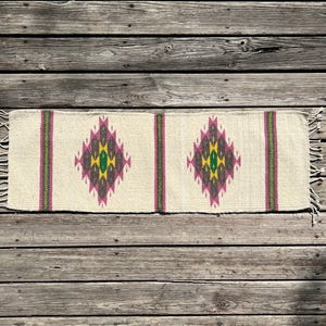 May include: A white woven rug with a geometric pattern in shades of pink, green, and yellow. The rug has a fringe on both ends.
