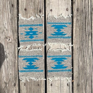 May include: Four gray woven coasters with a turquoise geometric design. Each coaster has a rectangular shape with a fringed edge. The coasters are displayed on a weathered wooden surface.