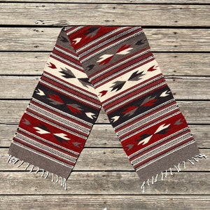 May include: A woven tapestry runner with a geometric pattern in shades of red, white, black, and gray. The runner has fringe on both ends.