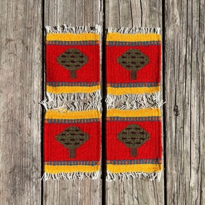 May include: Four woven coasters with a red background and a yellow border. Each coaster features a green tree design.