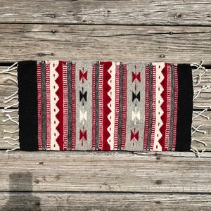 May include: A handwoven rug with a geometric pattern in shades of red, black, white, and gray. The rug has a fringe on both ends.