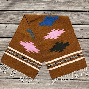 May include: A brown woven throw blanket with a geometric pattern in pink, blue, and black. The blanket has white fringe on the ends.