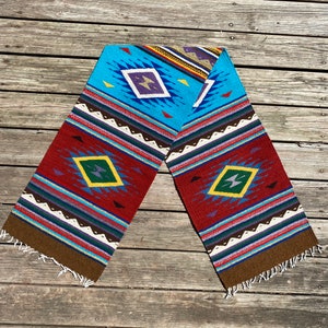 May include: A colorful woven table runner with a geometric pattern in shades of red, blue, brown, yellow, and green. The runner is folded in half and has fringed edges.