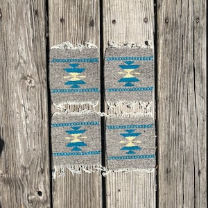 May include: Four gray woven coasters with a blue and yellow geometric pattern. The coasters are frayed on the edges and are sitting on a wooden surface.