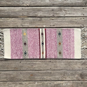 May include: A pink and white woven rug with a geometric pattern. The rug has a fringe on both ends.