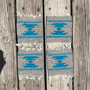 May include: Four woven coasters with a geometric pattern in shades of gray and blue. The coasters have a fringe border.