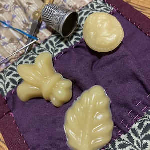 May include: Three small, beige, wax-like shapes on a purple fabric background. The shapes are a bee, a leaf, and a round shape with a raised design.