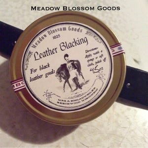 May include: A gold tin with a white label that reads "Meadow Blossom Goods 1825 Leather Blacking For black leather goods". The label also includes directions for use and a small illustration of a person riding a horse. The tin is 4.5 ounces and is made by Mrs. A. Schladweiler in Beechwood, Wisconsin.