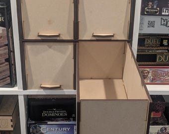 May include: A wooden game organizer with four closed compartments and one open compartment. The organizer is made of light brown wood with dark brown trim. The open compartment is on the bottom right, revealing the interior. The organizer is on a white shelf with board games.