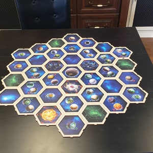 May include: A hexagonal game board with 36 tiles, each depicting a different planet or celestial body. The tiles are arranged in a honeycomb pattern, with a central tile featuring a large, dark planet. The board is made of wood and has a dark blue background.