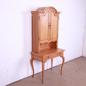 May include: A light wood desk with a hutch. The desk has two drawers and cabriole legs. The hutch has two doors and a shelf. The desk is in a white room.