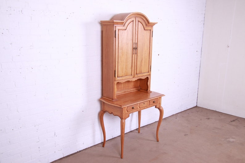 May include: A light wood desk with a hutch. The desk has two drawers and cabriole legs. The hutch has two doors and a shelf. The desk is in a white room.