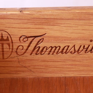 May include: Close-up of a wooden surface with a Thomasville logo. The logo features a crest with a crown and the word "Thomasville" in a cursive script.