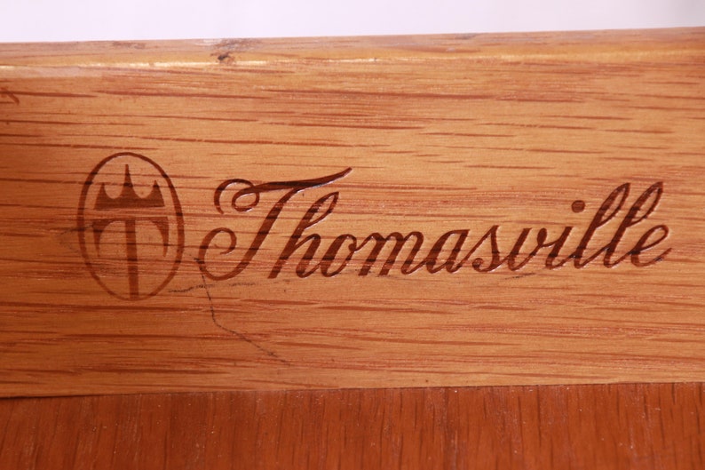 May include: Close-up of a wooden surface with a Thomasville logo. The logo features a crest with a crown and the word "Thomasville" in a cursive script.