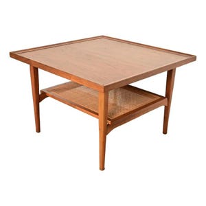 May include: A square wooden side table with a smooth, flat top surface and a raised edge. The table features four tapered legs and a lower shelf constructed with a woven cane mesh pattern, showcasing a natural medium brown wood grain finish.