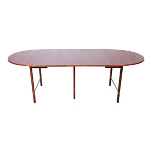 May include: A brown wooden oval extending dining table with a drop leaf on each side. The table has a simple design with four legs and a metal bar connecting the legs.