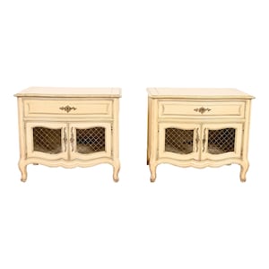 May include: A pair of off-white wooden nightstands with a single drawer and two doors with a wire mesh insert. Each nightstand has a curved bottom and four legs.