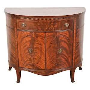 May include: A mahogany-colored, semi-circular cabinet with two doors and a drawer. The cabinet features a decorative wood grain pattern and brass hardware. The doors have oval inlays, and the drawer is centered above the doors. The cabinet has four curved legs.