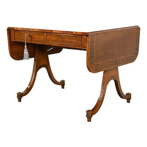 May include: A brown wooden drop-leaf table featuring two drawers with brass hardware and a decorative tassel hanging from the left drawer pull. The table is supported by four curved legs with fluted details at the base and a smooth top surface.