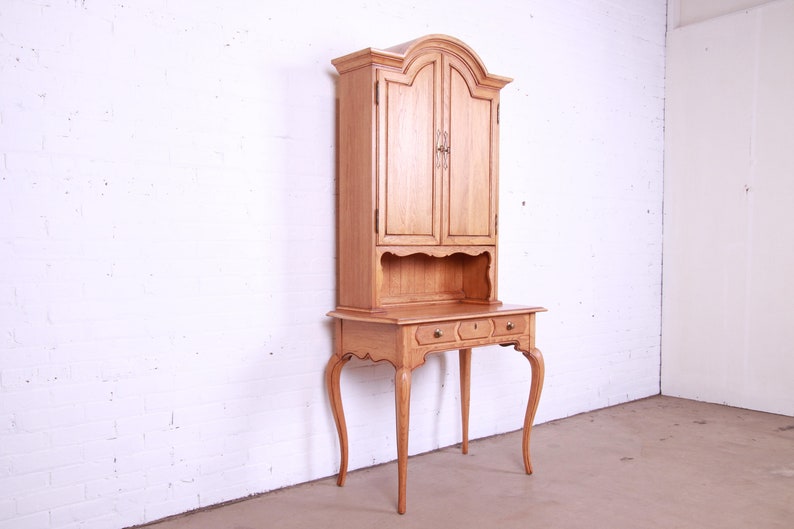 May include: A light wood desk with a hutch. The desk has a curved top and three drawers. The hutch has two doors and a shelf. The desk is in a white room.