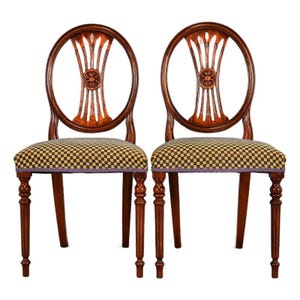May include: Two antique wooden chairs with a dark finish. The chairs have oval backs with a decorative carved design and checkered seat cushions. The legs are turned and have a decorative design. The chairs are set against a white background.