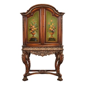 May include: A wooden cabinet with a decorative floral design. The cabinet has two doors with a green background and a floral painting. The cabinet sits on a carved base with ornate legs. The wood has a dark brown finish with gold accents.