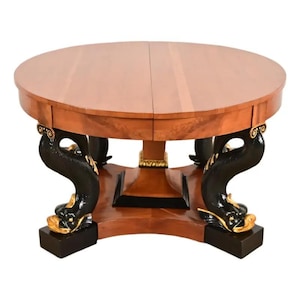 May include: A round wooden table with a light brown tabletop and dark brown legs. The legs are sculpted in a dolphin design with gold accents. The table has a pedestal base and is likely a dining table.