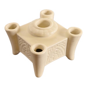 May include: A white ceramic candle holder with four candle slots and an ornate design. The holder has a square base and four legs.