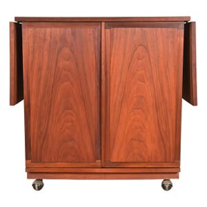 May include: A mid-century modern wooden cabinet with two doors and two drop-leaf extensions. The cabinet is made of reddish-brown wood and has a rectangular shape. It sits on small caster wheels.