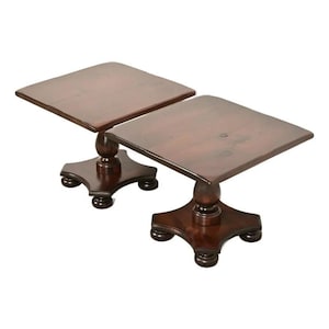 May include: Two dark brown wooden side tables. Each table has a square top, a turned pedestal base, and a four-footed base. The tables are made of a rich, dark wood and have a classic design.