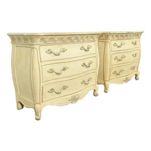 May include: Two cream-colored, antique-style dressers with three drawers each. The dressers have curved fronts, ornate silver-toned handles, and decorative trim along the top and bottom. The legs are curved and end in small feet.