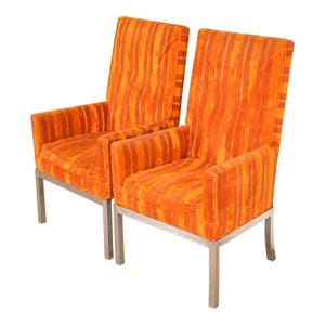 May include: Two orange armchairs with vertical striped patterns. The chairs have a high back and armrests, with a silver metal frame. The upholstery is a vibrant orange color, creating a bold statement.