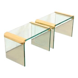 May include: Three clear glass tables with rectangular tops and curved, gold-colored metal accents. The tables are arranged in a staggered formation, showcasing their minimalist design and modern aesthetic. The glass is transparent, allowing light to pass through.