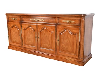 May include: A long, wooden sideboard with four doors and three drawers. The doors have a raised, arched design and the drawers have ornate brass handles. The sideboard is made of a light wood with a warm, honey-colored finish.