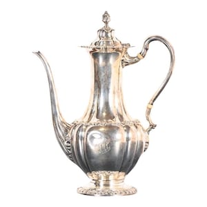 May include: A silver coffee pot with a fluted body and a decorative lid. The pot has a handle and a spout. The pot is on a white background.